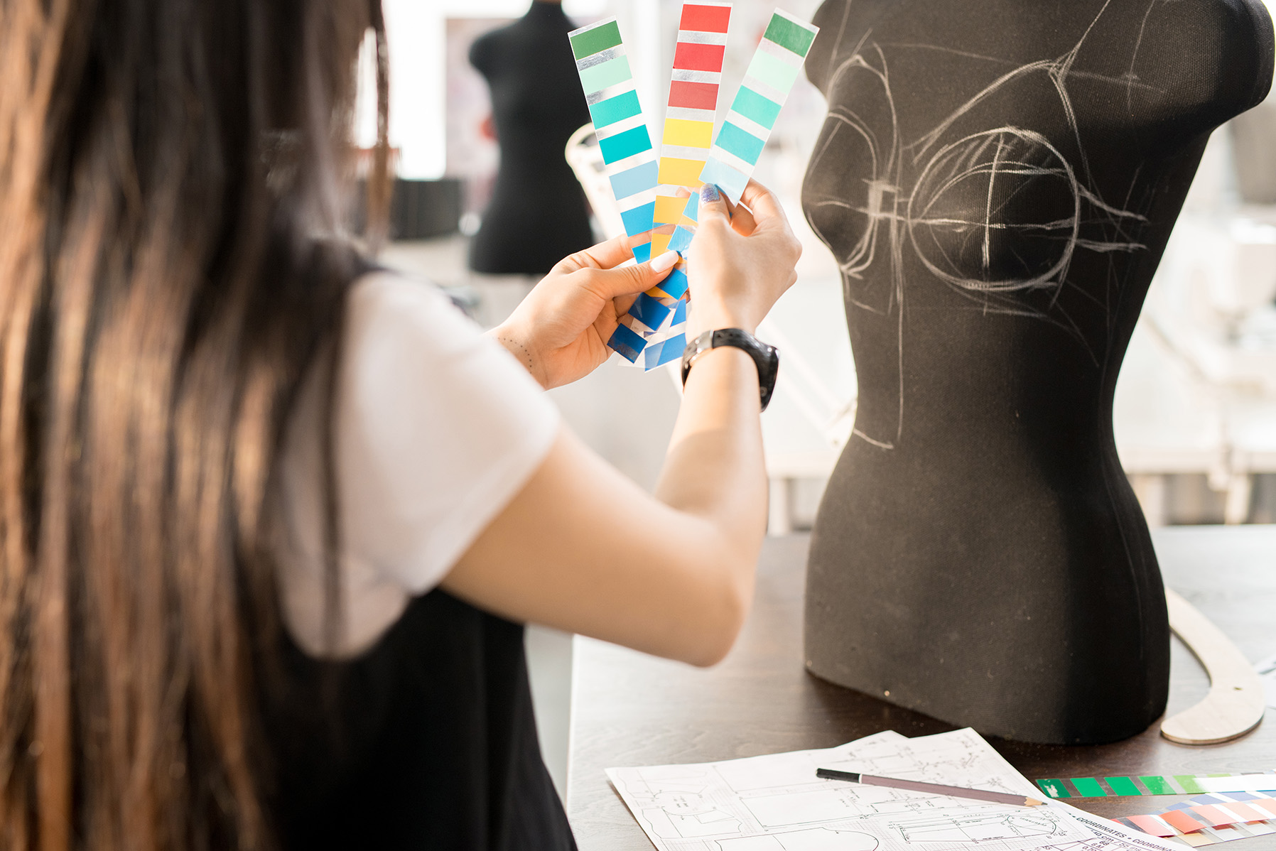 Back view portrait of focused Asian woman working in fashion design choosing color palette holding swatches standing at table by sewing dummy in modern sunlit atelier studio.